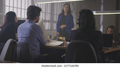 Group Of Young Office Workers Gathered Together For A Meeting At Night. Male Employee Sharing Information With Female Co Worker. Employees Sitting At Table In Conversation