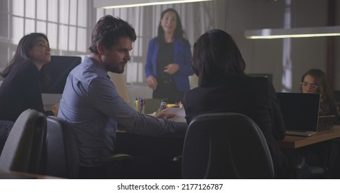 Group Of Young Office Workers Gathered Together For A Meeting At Night. Male Employee Sharing Information With Female Co Worker. Employees Sitting At Table In Conversation