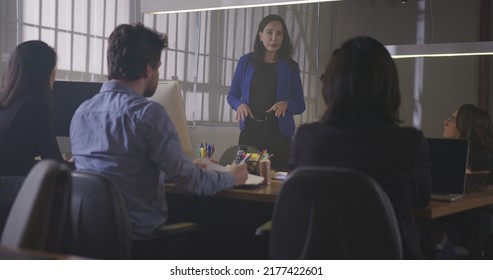 Group Of Young Office Workers Gathered Together For A Meeting At Night. Male Employee Sharing Information With Female Co Worker. Employees Sitting At Table In Conversation