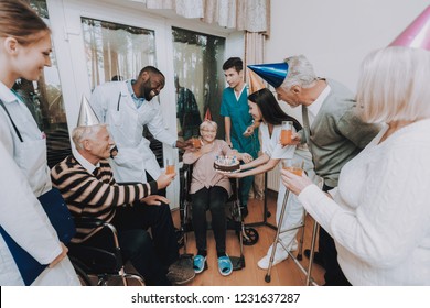 Group Young And Old People Congratulate Woman. Birthday In Nursing Home. Elderly Female Very Surprised. Woman Very Happy. Guests Hold Glasses Of Drinks. Nurse Hold Cake. Birthday Party Celebration.