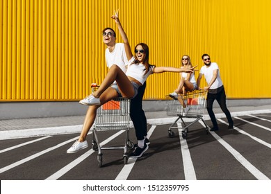 A Group Of Young People, Friends In White T-shirts, Fun Ride On Carts Near The Store, Supermarket, Friends Having Fun, Laughing In Sunny Weather. Shopping, Discounts, Outdoors