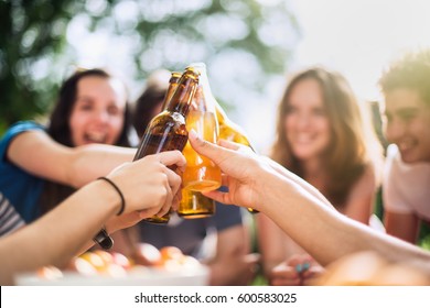 Group Of Young People Having Fun Sitting Around A Table Outside. Clinking Their Bottles Of Beer, Focus On The Bottles. Shot With Flare