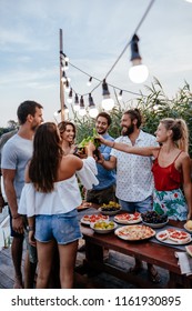 Group Of Young People Having Outdoor Dinner Party By The River And Rising Bottles.