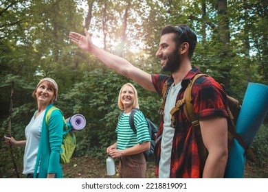 Group Of Young People Hikers Walking In The Woods. Man Showing To Friends The Way For Hiking Trail. Backpackers In Sunny Forest