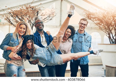 Group of young people holding their friend girl on hands in front of them posing to camera and laughing, standing indoors in warm light