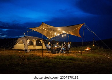 Group of young people out for the weekend eating BBQ after sunset under a canvas roof while camping on a lakeside park vacation. - Powered by Shutterstock