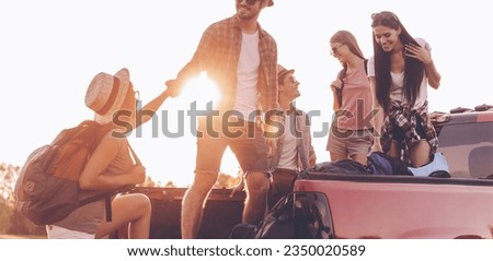 Group of young people preparing to road trip while man helping woman to get to pick-up truck