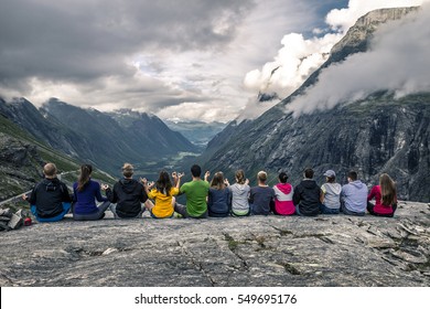 Group Of Young People Sitting On The Plateau On Top Of Mountain And Looking Forward On The Beautiful Valley Full Of Clouds, Trollstigen, Norway