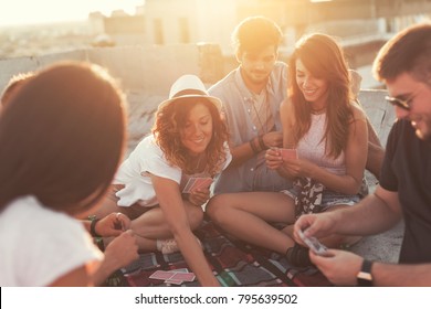 Group of young people sitting on a picnic blanket, having fun while playing cards on the building rooftop. Focus on the couple in the middle
