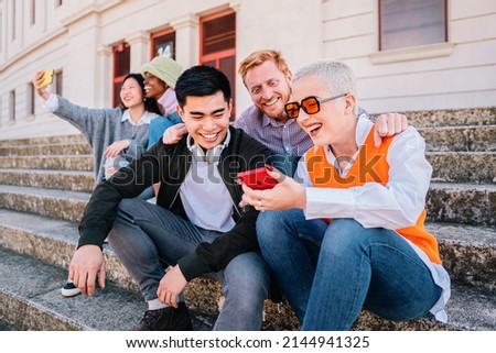 Group of young people sitting at outdoor watching socialmedia in the smartphone. Happy multiethnic hipsters looking funny online content on mobile phone.