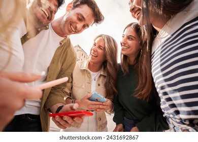 A group of young people are standing in a circle, smiling and sharing a cell phone. Happy friends using mobile laughing and having fun togethe - Powered by Shutterstock