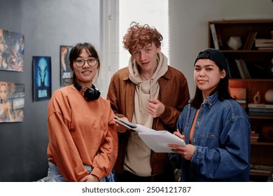 Group of young people standing together in art studio holding documents One person wearing headphones, another lifting hand, creating sense of teamwork and collaboration in studio - Powered by Shutterstock