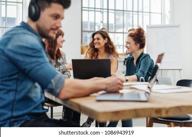 Group of young people working or studying together around a wooden table in a bright airy room with focus to a three girls in the background chatting and smiling - Shutterstock ID 1086943919