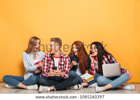 Group of young positive school friends doing homework while sitting on a floor together with laptop computers over yellow background