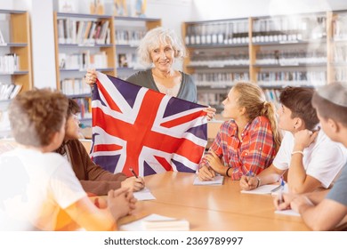 Group of young pupils listening to female teacher holding flag of the Great Britain in school library - Shutterstock ID 2369789997