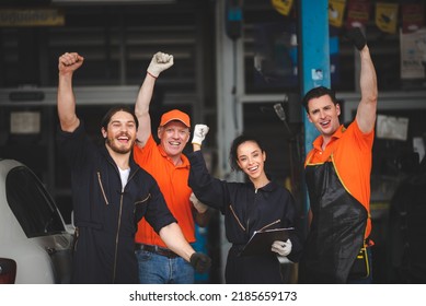 Group of young and senior male and female car mechanics wearing uniform with cap and gloves in garage enjoying and celebrating with dance and smiling with hands up while holding clipping board - Powered by Shutterstock