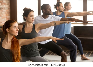 Group of young sporty afro american and caucasian people practicing yoga lesson, standing in Warrior two exercise, Virabhadrasana II pose, working out, indoor close up, studio background - Powered by Shutterstock