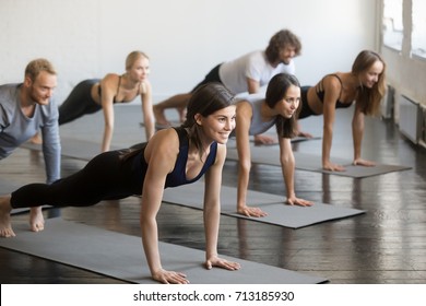 Group of young sporty attractive people practicing yoga lesson with instructor, doing Push ups or press ups exercise, standing in Plank pose, friends working out in club, indoor full length, studio  - Powered by Shutterstock