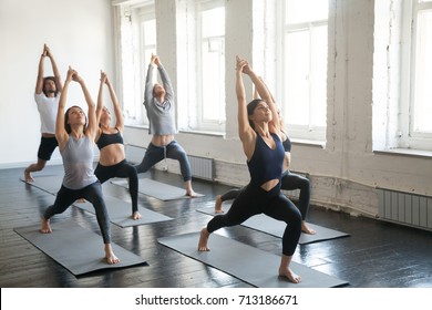 Group of young sporty attractive people practicing yoga lesson with instructor, standing together in Virabhadrasana 1 exercise, Warrior one pose, working out, indoor full length, studio background 