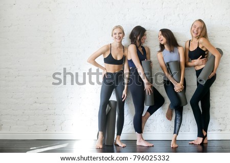 Group of young sporty girls with yoga mats standing at white wall. Female companions in gym resting after fitness, indoor full length, loft studio, copy space
