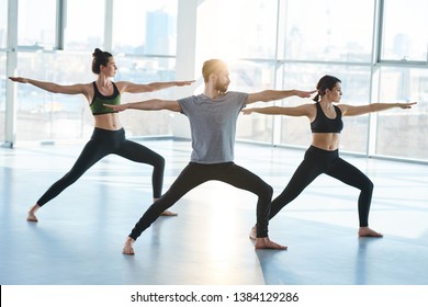 Group of young sporty people in activewear standingon the floor in one of yoga positions with their arms outstretched