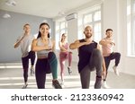 Group of young sporty people doing exercises together in bright and spacious gym. Happy men and women doing aerobics exercises lifting high knees in gym. Fitness, sport, aerobics and people concept.