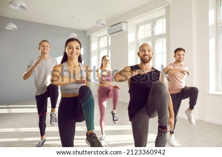 Group of young sporty people doing exercises together in bright and spacious gym. Happy men and women doing aerobics exercises lifting high knees in gym. Fitness, sport, aerobics and people concept.