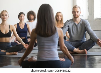 Group Of Young Sporty People Practicing Yoga Lesson With Instructor, Doing Padmasana Exercise, Lotus Pose, Students Working Out In Studio, Teacher Rear View. Healthy, Mindful Lifestyle Concepts