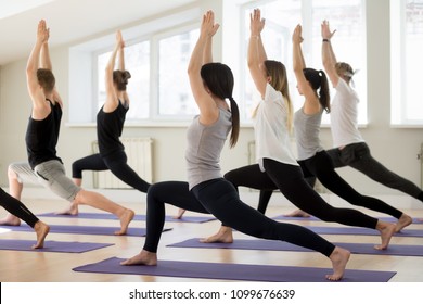 Group of young sporty people practicing yoga lesson, doing Warrior I exercise, Virabhadrasana 1 pose, working out, indoor full length, yogi students training in sport club, studio session. Back view