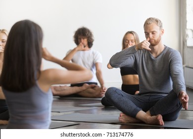 Group Of Young Sporty People Practicing Yoga Lesson With Instructor, Sitting, Making Alternate Nostril Breathing, Nadi Shodhana Pranayama Exercise, Sukhasana Pose, Working Out, Indoor Full Length 