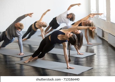 Group of young sporty people practicing yoga lesson with instructor, stretching in Bending Side Plank exercise, Vasisthasana pose, working out, indoor studio image. Wellbeing, wellness concept 
