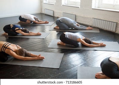 Group Of Young Sporty People Practicing Yoga Lesson With Instructor, Lying In Balasana Exercise, Child Pose, Friends Working Out In Club, Indoor Full Length, Studio. Wellbeing, Wellness Concept 