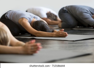 Group of young sporty people practicing yoga lesson with instructor, sitting in Balasana exercise, Child pose, friends working out in club, indoor close up image, studio. Wellbeing, wellness concept 