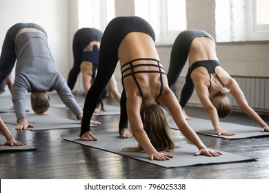 Group of young sporty people practicing yoga lesson with instructor, stretching in Downward facing dog exercise, adho mukha svanasana pose, working out, indoor full length, students training, studio