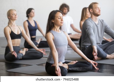 Group of young sporty people practicing yoga lesson with instructor, sitting in Padmasana exercise, Lotus pose, working out, indoor full length, students training in club, studio