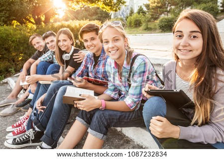 group of young students with books and gadgets sit on the steps in the park