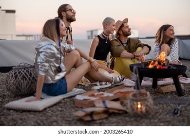 Group of young stylish friends sitting together by the fireplace, having a picnic on the rooftop terrace at dusk. Enjoy summertime and beautiful sunset view