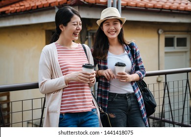 Group Of Young Stylish Happy People Leaning Against Railing And Talking While Drinks Coffee In Paper Cup Standing Outdoor In Small Town With Red Brick Roof Of Old Historic House. Two Girls Laughing