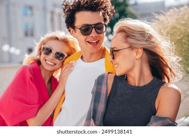 Group of young three stylish friends posing in the street. Fashion man and two cute female dressed in casual summer clothes. Smiling models having fun. Cheerful women and guy outdoors, in sunglasses