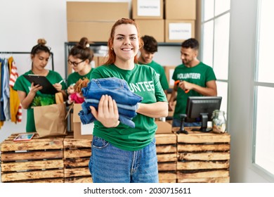 Group of young volunteers working at charity center. Woman smiling happy and holding stack of folded jeans to donate.