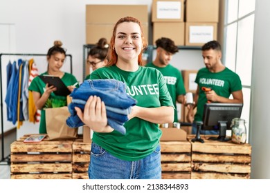 Group of young volunteers working at charity center. Woman smiling happy and holding stack of folded jeans to donate.