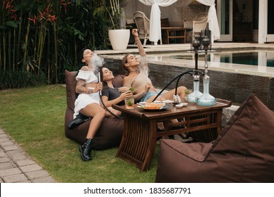 Group of young women with dark hair smoking a hookah or shisha, exhaling white smoke on a bean bags. Evening rest in an outdoor sushi bar. Summer. Vacation.