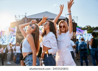 Group of young women enjoying at a beach music festival, dancing together in summer outfits with sunglasses. Sunlight illuminates the happy crowd, friends celebrate, party atmosphere by the ocean. - Powered by Shutterstock