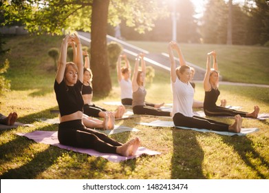 Group of young women are practicing yoga and raising hands above heads morning in park while sunrise. Group of people are sits outdoors on grass with eyes closed