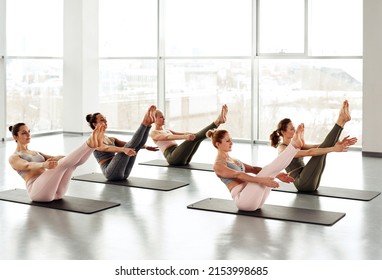 Group of young women sitting on yoga mats and doing boat pose in modern studio with panoramic windows - Powered by Shutterstock