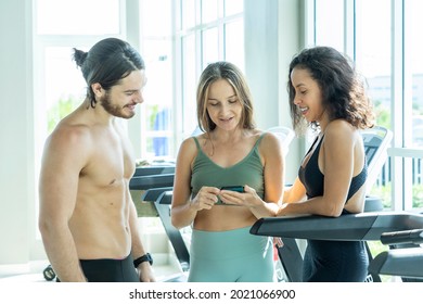 Group Of Young Women In Sportswear Holding Smartphones To Evaluate Exercise Results. Male Trainer Talking To A Latin American Woman  Discussing Exercise At The Gym. 