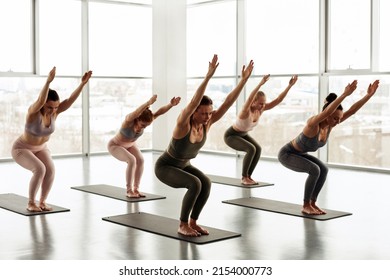 Group Of Young Women Standing On Mats And Performing Chair Pose With Raised Arms In Yoga Studio