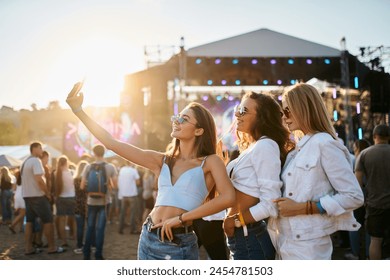 Group of young women take selfie at summer beach music festival. Happy friends party, dance in crowd. Sun sets behind stage. Girls enjoy live concert, fun vacation. Casual fashion, youth culture. - Powered by Shutterstock