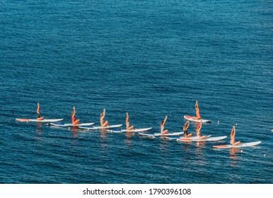 Group of young womens in swimsuits doing yoga on sup board in calm sea, early morning. Balanced pose - concept of healthy life and natural balance between body and mental development.