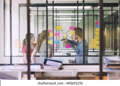 Groupe Of Casual Business People Working In Front Of Glass Wall Using Notes Papers Post It. Seen Through Glass In Startup Office.